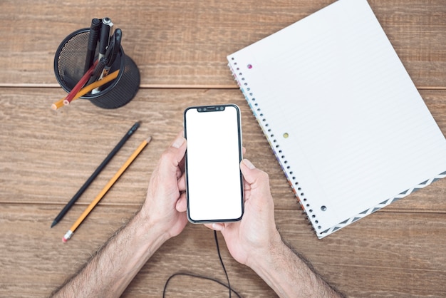 Man's hand using mobile phone on wooden desk with stationeries and spiral notebook