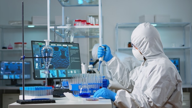 近代的な設備の整った実験室でマイクロピペットを使用してつなぎ服を着た男性科学者。 covid19ウイルスの開発研究のためのハイテクおよび化学ツールを使用してワクチンの進化を調べる医師のチーム