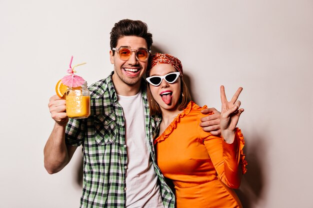 Man in shirt and glasses holds cocktail and hugs his beloved girlfriend. Woman in orange dress, headscarf and glasses shows tongue and peace sign.