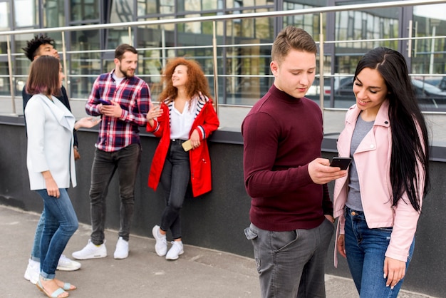 Man showing smartphone to his female friend standing near their group of friends