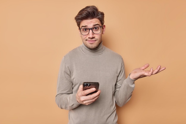 Бесплатное фото Мужчина пожимает плечами, носит очки и водолазку держит мобильный телефон читает что-то странное, просматривает ленту новостей в социальных сетях, недоумевает по поводу онлайн-контента