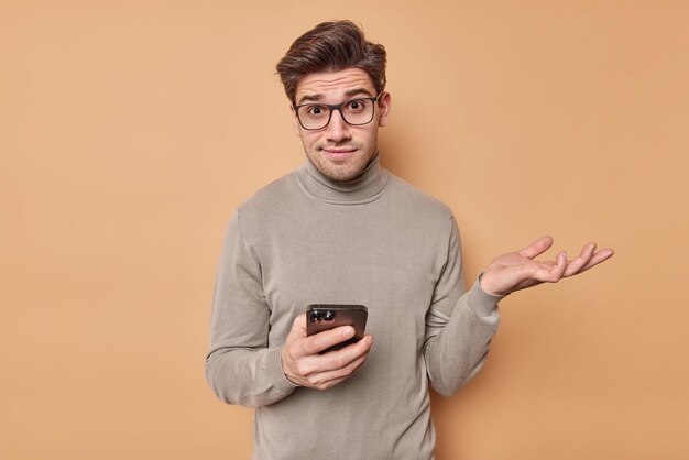 man shrugs shoulders wears optical glasses and turtleneck holds mobile phone reads something strange browses newsfeed via social networks puzzled about online content