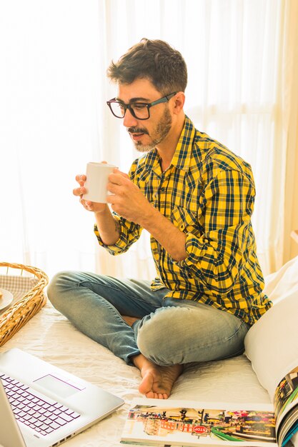 Foto gratuita equipaggi la seduta sul letto che beve il caffè che esamina il computer portatile