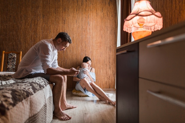 Free photo man sitting on bed using mobile phone with his wife carrying her baby