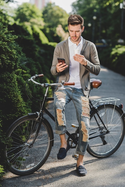 Man sitting on bicycle holing disposable coffee cup using cell phone