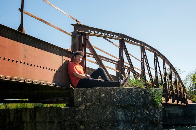 Foto gratuita uomo che si siede sul pilastro del ponte che guarda l'obbiettivo