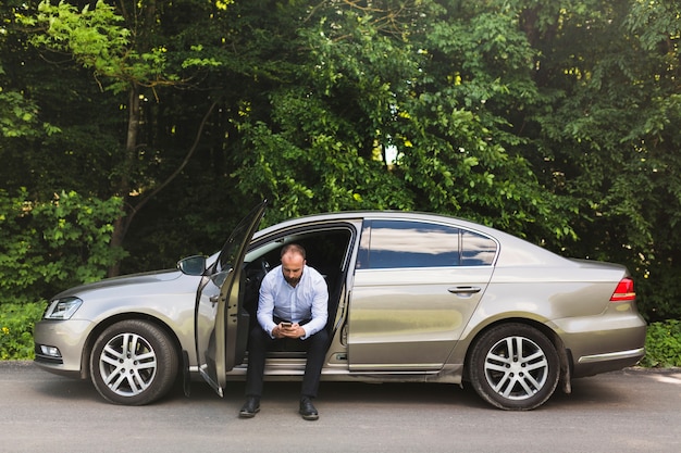 Free photo man sitting in a car with open door using mobile phone