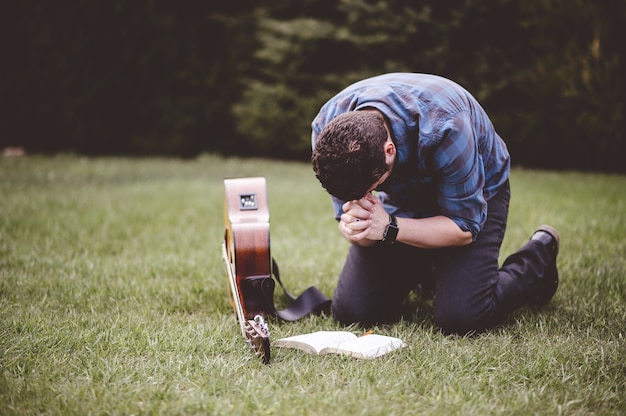 Uomo seduto sull'erba e pregando con un libro e una chitarra vicino a lui