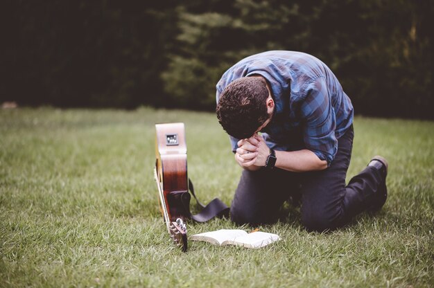 Человек сидит на траве и молится с книгой и гитарой рядом с ним
