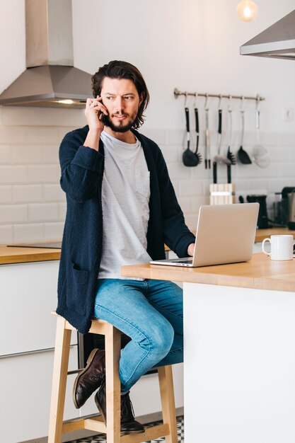 Man sitting in kitchen using laptop and talking on cell phone looking away