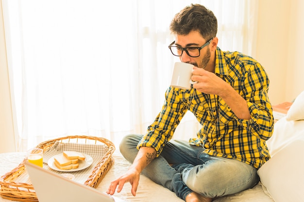 朝食とノートパソコンをベッドの上でコーヒーを飲みながらベッドの上に座っている男