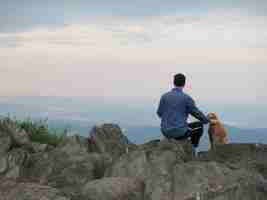 무료 사진 바위에 앉아 흐린 하늘 아래 산으로 둘러싸인 개를 쓰다듬는 남자