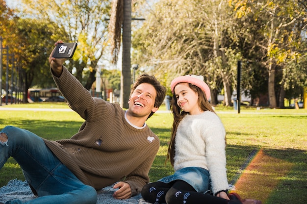 Человек, сидящий в парке с его дочерью, берущий себя с помощью смартфона