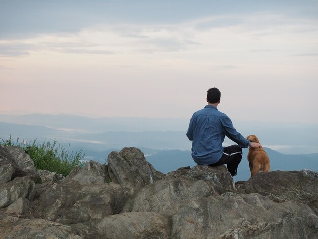 Man sitting on the rock and petting a dog surrounded by mountains under a cloudy sky