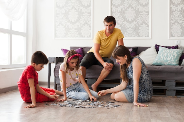 Man sitting on sofa and looking at his wife and children playing jigsaw puzzle at home