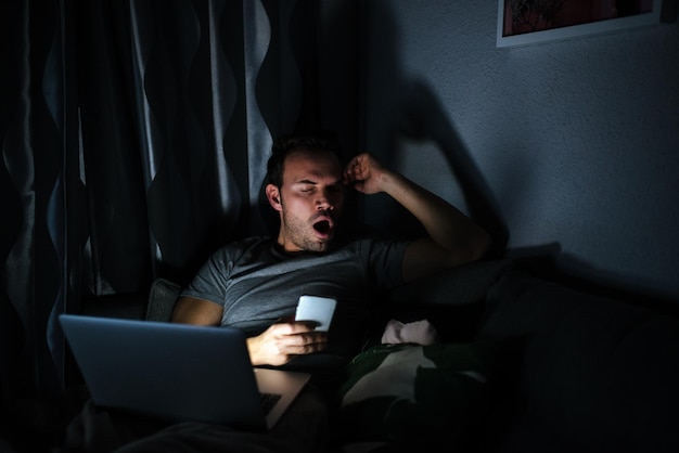 Man sitting on the sofa with a mobile phone and computer yawing