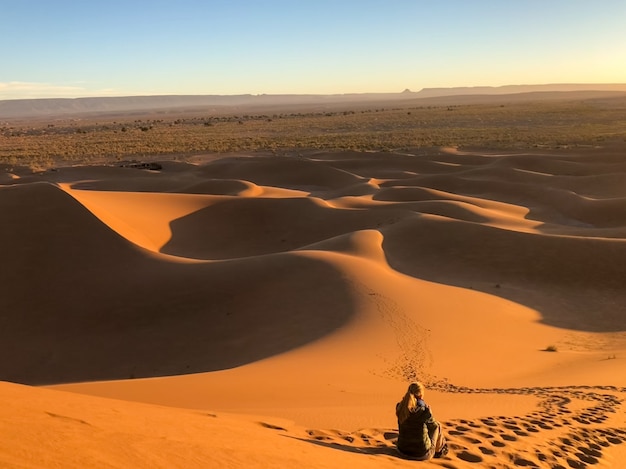 Man sitting on sun dunes at a desert surrounded by tracks