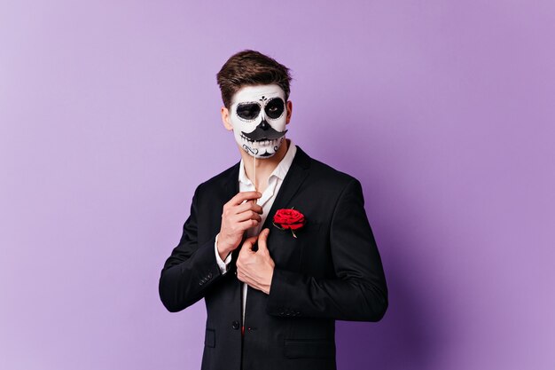 Man in skull mask winks coquettishly, posing with overlaid mustache for portrait on isolated background.