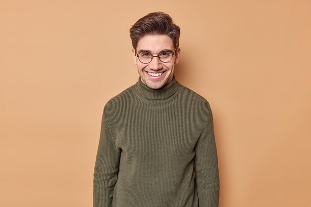 man smiles gladfully with teeth expresses cheerful emotions listens funny story from interlocutor wears round spectacles and jumper isolated on beige wall.