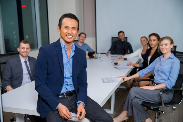 Man smiling with suit sitting at a table with colleagues behind