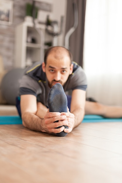 Uomo in abbigliamento sportivo che allunga le gambe sul tappetino da yoga durante la pandemia globale.