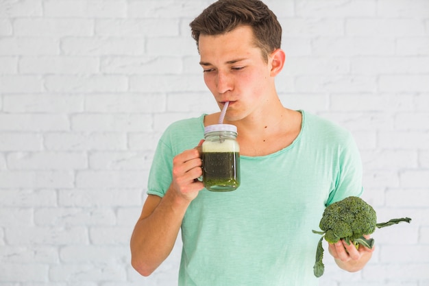 Foto gratuita equipaggi la condizione contro il muro di mattoni bianco che beve il frullato fresco dei broccoli in barattolo