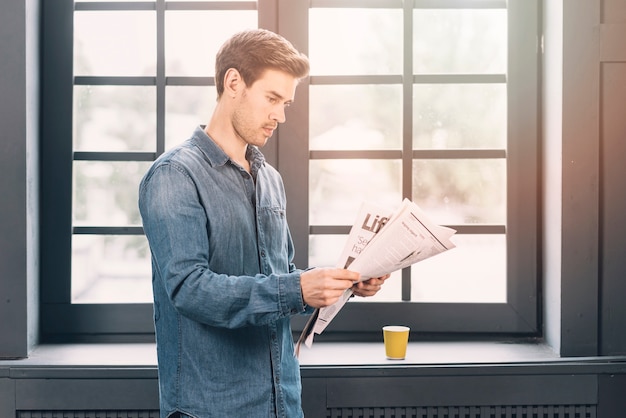 A man standing near the closed window reading newspaper