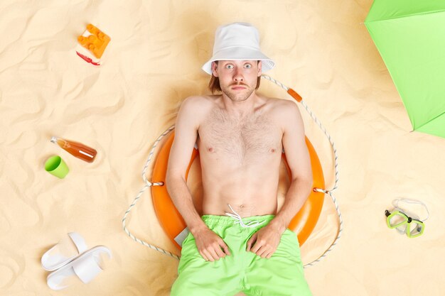  man stares at camera with bugged eyes cannot believe in something amazing wears white panama green shorts lies on inflated lifebuoy spends free time at beach has lazy day