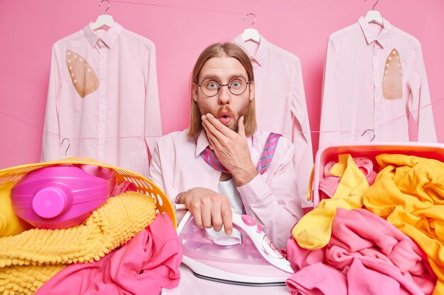 man stares shocked irons clothes surrounded by stacks of laundry poses on pink