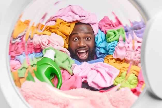  man sticks head through multicolored laundry poses through washing machine drum with bottle of detergent