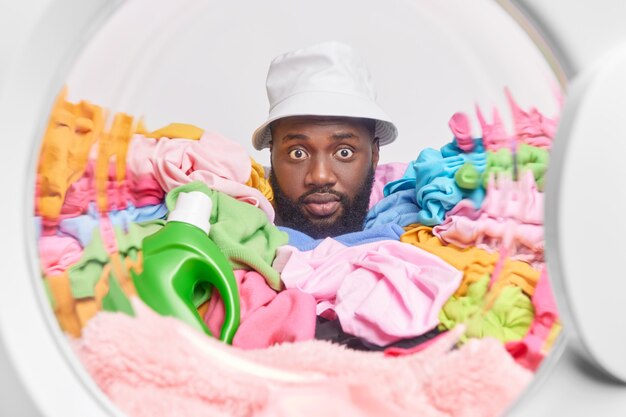  man sticks head in washing machine door poses around colorful laundry with bottle of detergent wears panama busy doing washing. Washer full of dirty clothing