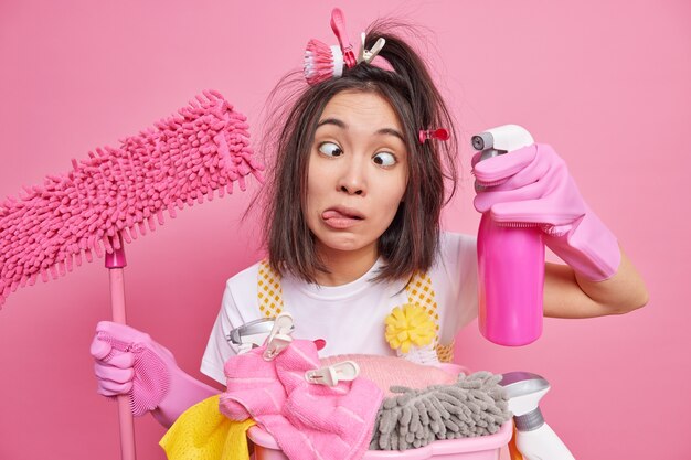 man sticks out tongue crosses eyes makes grimace foolishes around while cleaning house holds cleaning detergent and mop stands near stack of laundry poses on pink