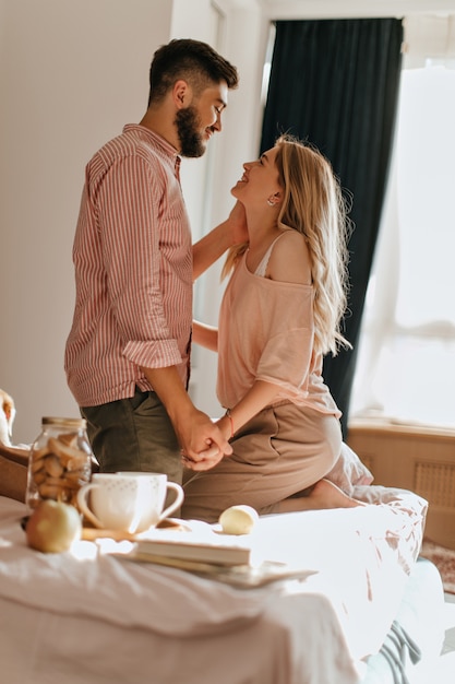 Man in striped shirt gently holds hand of his girlfriend, sitting on white bed during breakfast with tea and cookies.