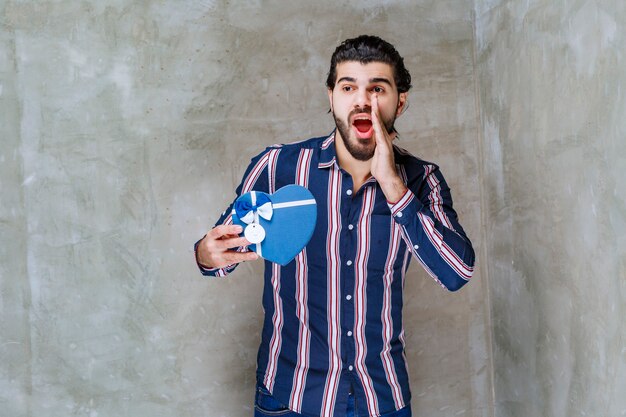 Man in striped shirt holding a blue heart shape gift box and calling someone to come nearby