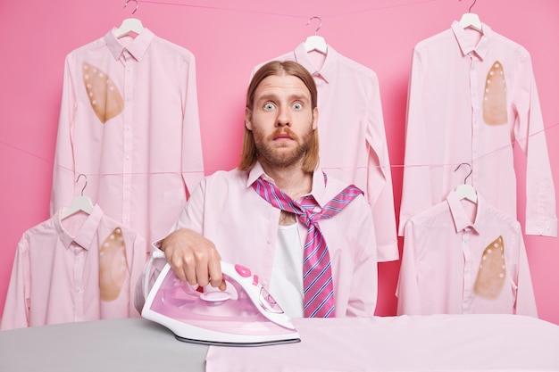 man strokes clothes uses electric stream iron wears shirt and tie around neck has much work to do poses on pink 