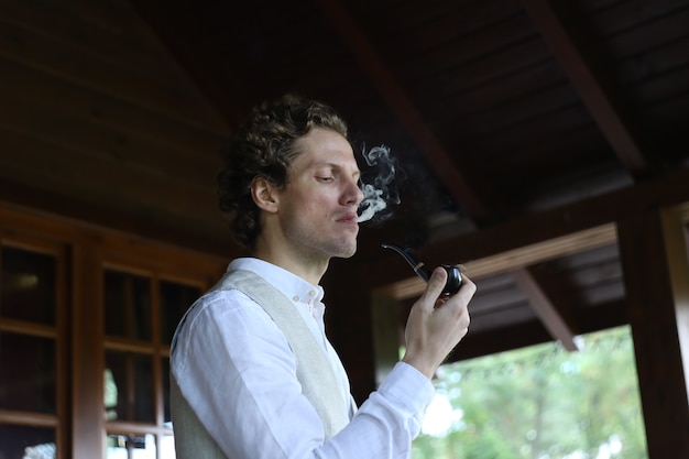 Foto gratuita uomo in abiti eleganti che fuma una pipa rilasciando fumo all'esterno della residenza