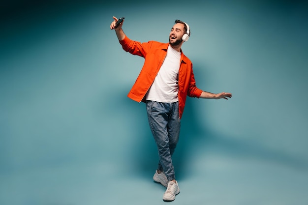 Man in stylish outfit dancing on blue background and holding smartphone