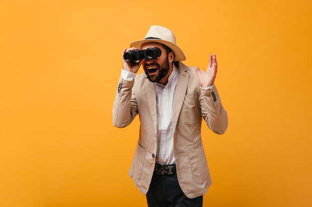 Man in suit and hat posing with binoculars on orange background