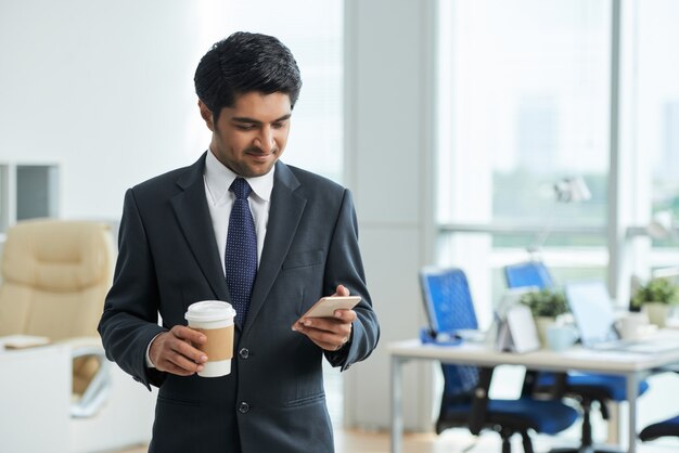 Man in suit standing in office, holding takeaway coffee and using smartphone