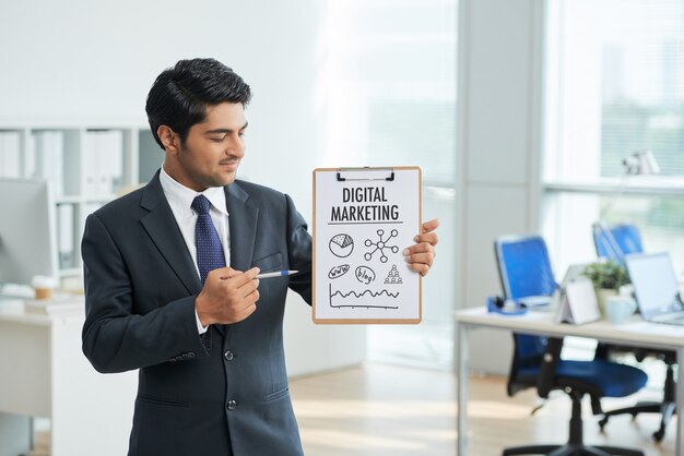 Man in suit standing in office with clipboard and pointing to poster with words 