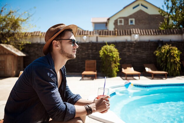 Man in sunglasses and hat drinking cocktail, sitting near pool