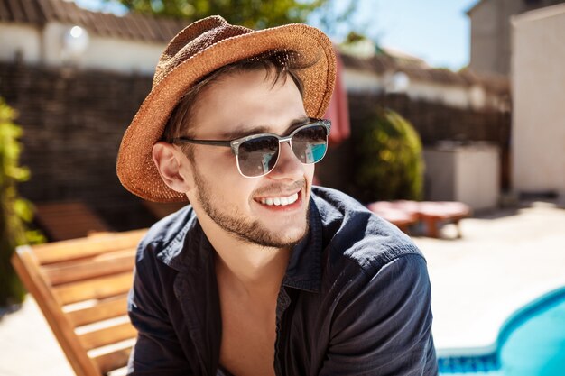 Man in sunglasses and hat smiling, sitting near swimming pool