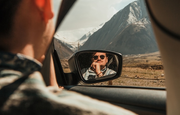 man in sunglasses looking at the mountains with car window reflected in the mirror