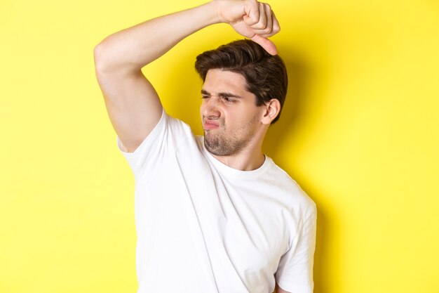 Man in sweat smelling his armpit, standing in white t-shirt and grimacing from stinky clothes.