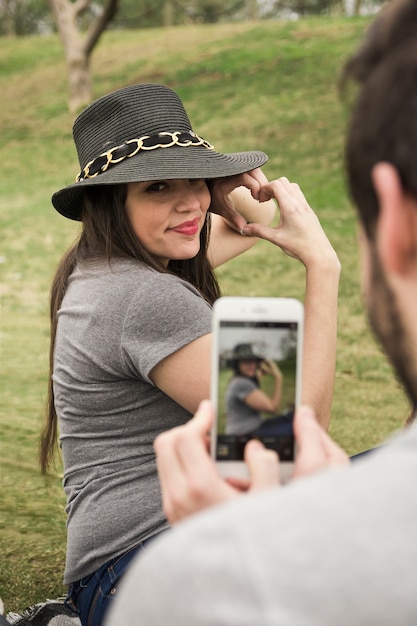 Man taking photo of her girlfriend making heart with hand from cell phone