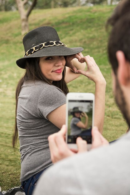 Man taking photo of her girlfriend making heart with hand from cell phone