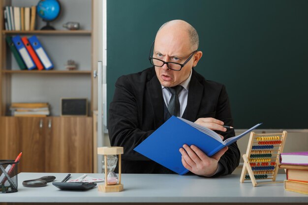Man teacher wearing glasses checking class register looking aside being displeased making wry mouth sitting at school desk in front of blackboard in classroom