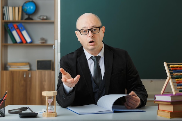 Man teacher wearing glasses checking class register looking at camera being confused raising arm in displeasure sitting at school desk in front of blackboard in classroom