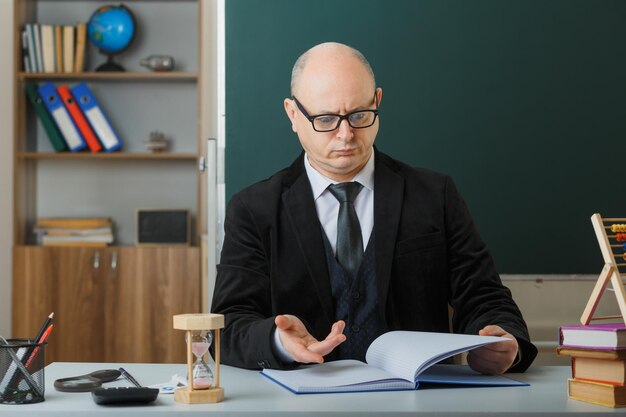 Man teacher wearing glasses checking class register looking confused and displeased raising arm in displeasure sitting at school desk in front of blackboard in classroom