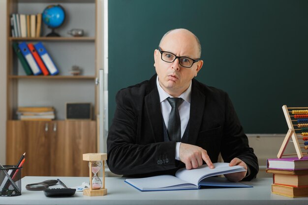 Учитель-мужчина в очках, проверяющий школьный журнал, выглядит смущенным и удивленным, сидя за школьной партой перед доской в классе